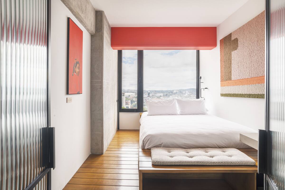 A modern bedroom with a large window, a bed, a bench, and a red accent wall.