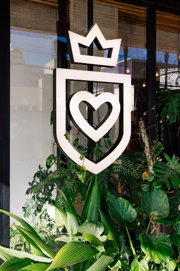 A logo with a crown and heart on a glass window, surrounded by green plants.