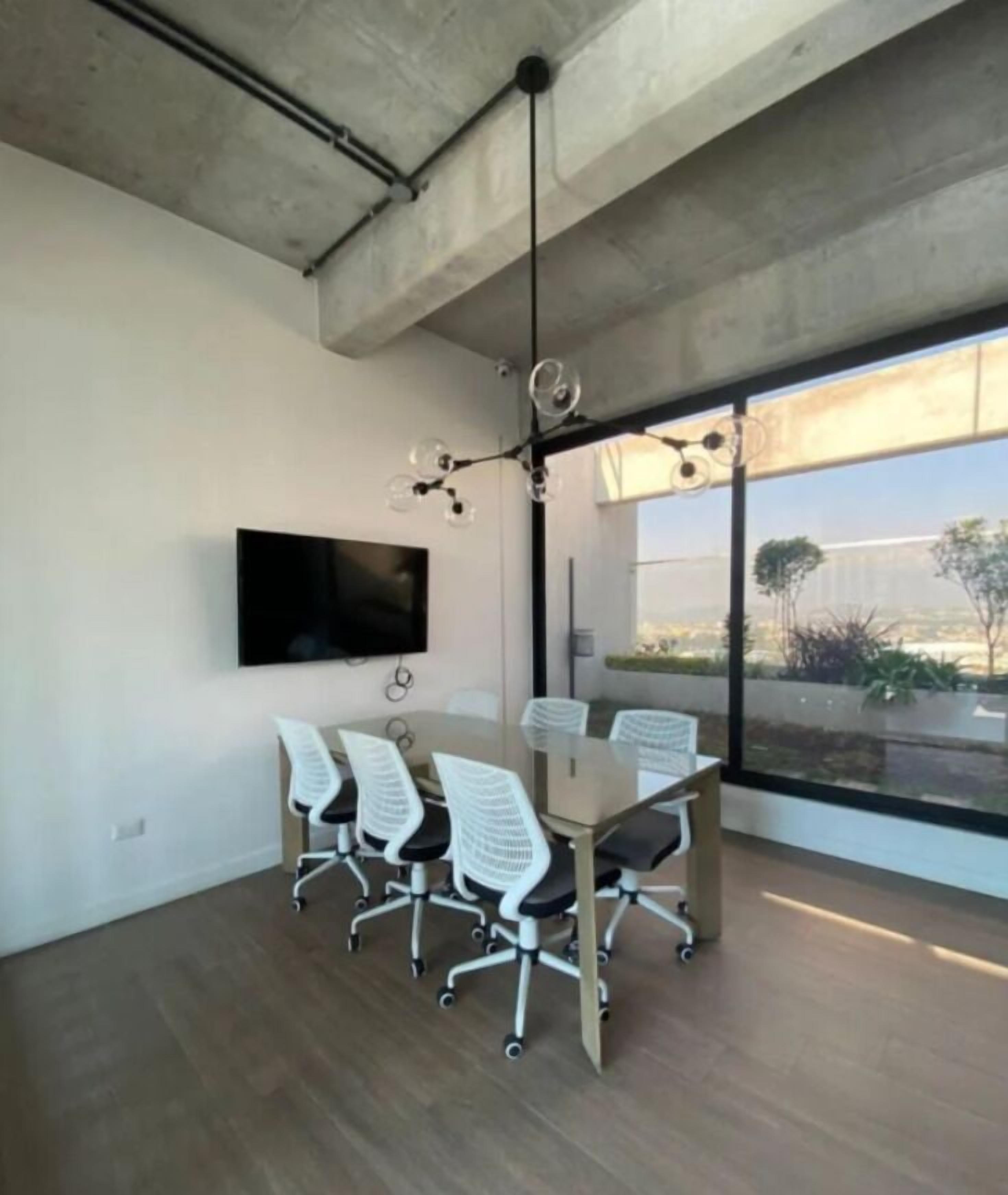 A modern meeting room with a large table, six office chairs, a flat-screen TV, and a large window offering a view of the outdoors.