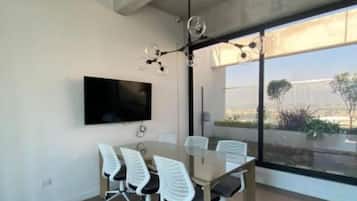 A modern meeting room with a large table, six office chairs, a flat-screen TV, and a large window offering a view of the outdoors.