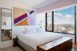 Ein modernes Schlafzimmer mit einem großen Bett, Blick auf die Stadt und einem farbenfrohen, abstrakten Gemälde an der Wand.