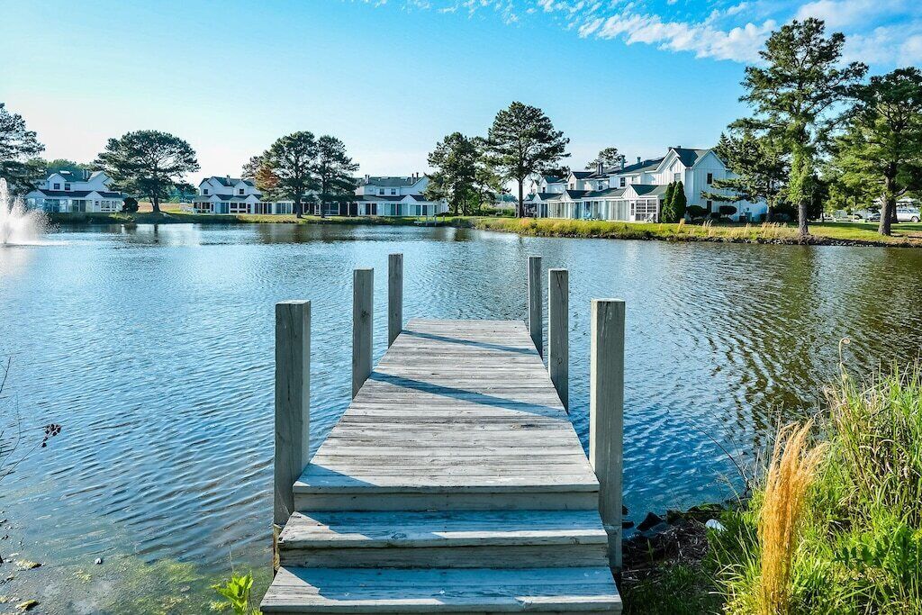 A wooden pier leading to a calm lake with houses in the background.