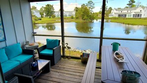 Eine Terrasse mit einer Couch und Stühlen, einem Tisch mit einem Eimer und Blick auf einen See mit Häusern im Hintergrund.