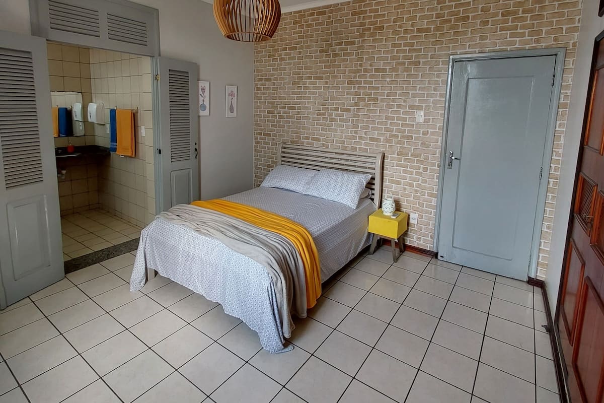 Um quarto com cama, parede de tijolos, porta e chão ladrilhado.
