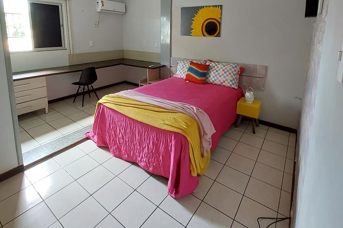Quarto com cama, escrivaninha, cadeira, janela e aparelho de ar condicionado.