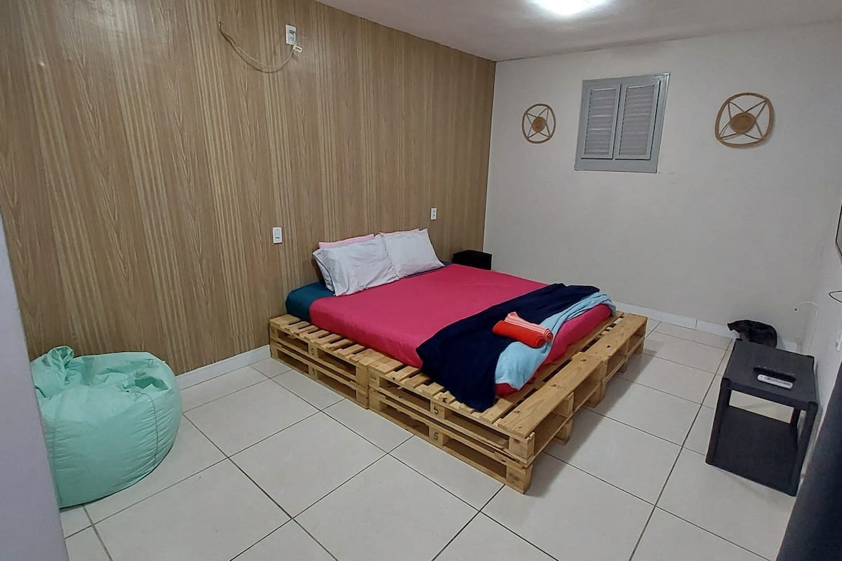 Quarto com cama sobre palete de madeira, uma poltrona de grãos, uma mesinha e prateleira fixada na parede.