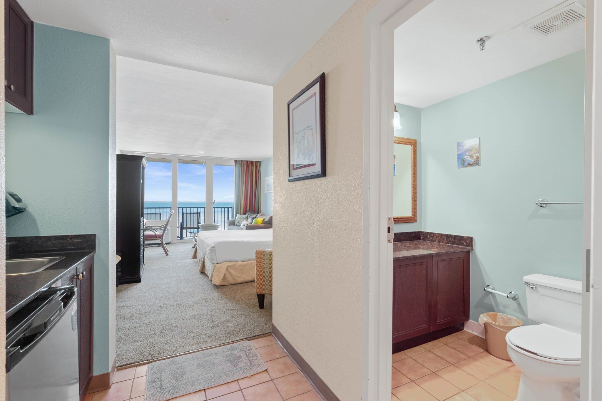 A hotel room with a kitchenette, a bed, a balcony with a view, and a bathroom.