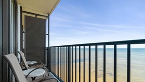 Ein Balkon mit zwei Stühlen und einem Tisch, der auf einen Strand mit klarem Wasser und einem Metallgeländer blickt.