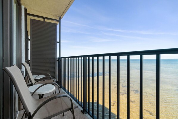 A balcony with two chairs and a table, overlooking a beach with clear water and a metal railing.