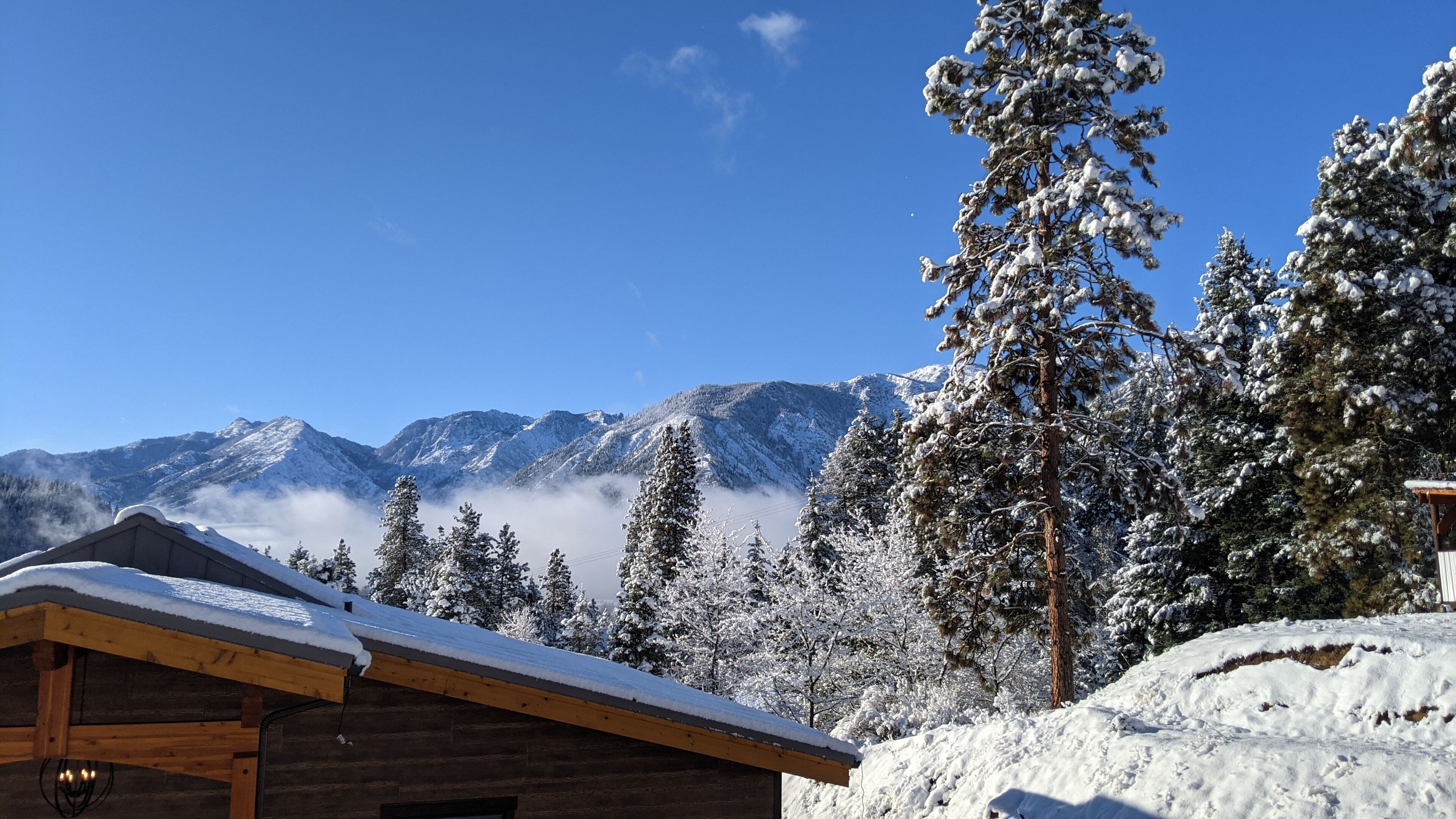 Une cabane en bois, dont le toit est recouvert de neige, entourée d’arbres chargés de neige et avec des montagnes en arrière-plan.