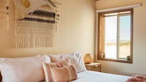 A bed with white and patterned pillows, a macrame wall hanging, a small table with a lamp, and a window with a view of the beach. - The Magic Hour House | Spa | Pool | Games | Cozy (Joshua Tree)