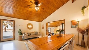 A spacious room with a wooden ceiling, a large wooden dining table, white chairs, a sofa, and a large mirror. - The Magic Hour House | Spa | Pool | Games | Cozy (Joshua Tree)