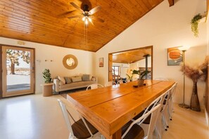 A spacious room with a wooden ceiling, a large wooden dining table, white chairs, a sofa, and a large mirror. - The Magic Hour House | Spa | Pool | Games | Cozy (Joshua Tree)