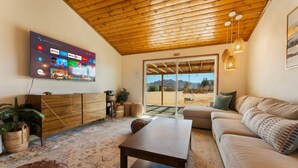 A living room with a wooden ceiling, a flat-screen TV, a sofa, a coffee table, and a view through a glass door. - The Magic Hour House | Spa | Pool | Games | Cozy (Joshua Tree)