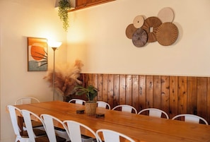 Zona pranzo con tavolo di legno e sedie bianche, un pannello decorativo a parete e una pianta pensile.
