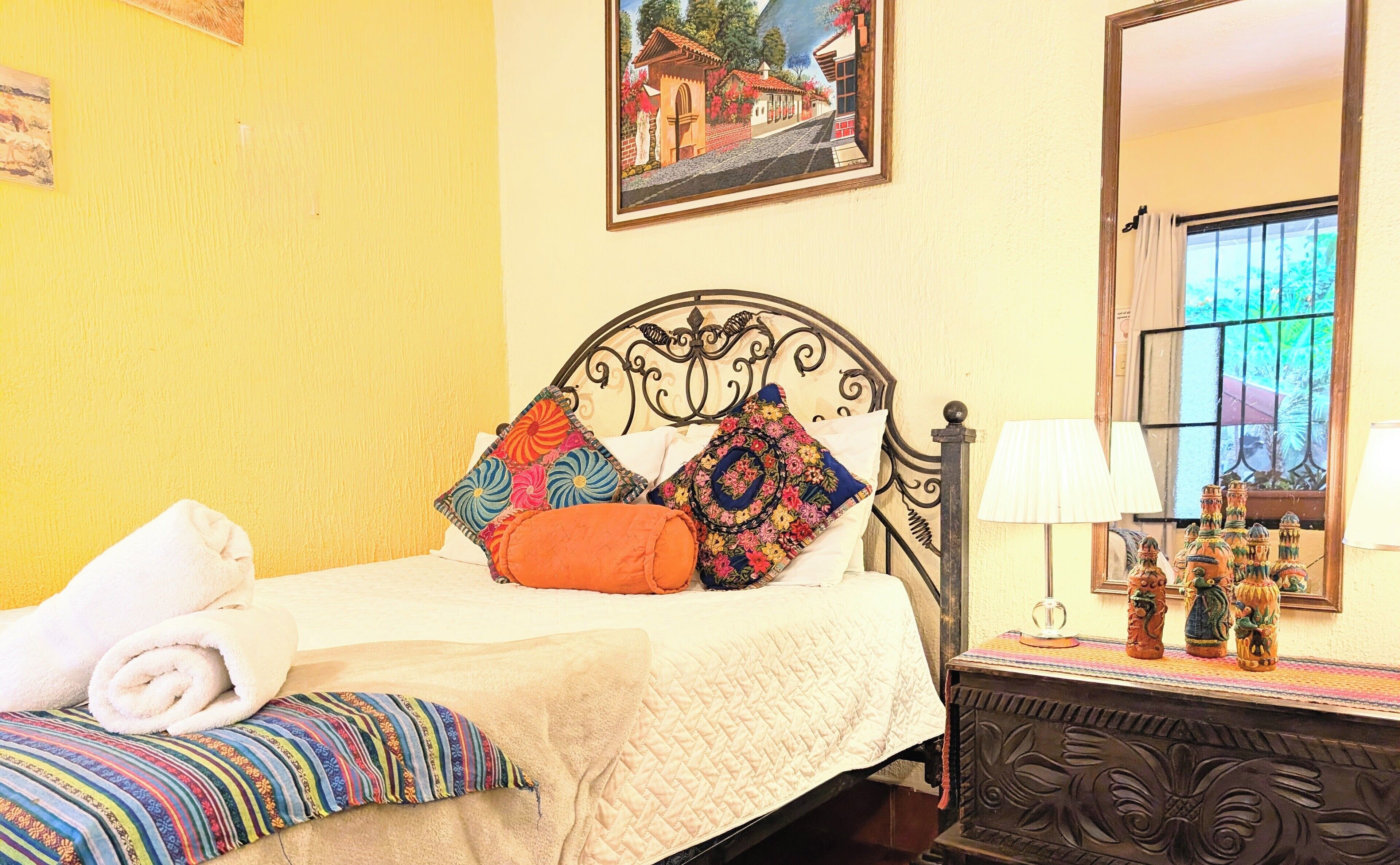 A neatly made bed with colorful pillows, a window with bars, and a decorative vase on a nightstand.