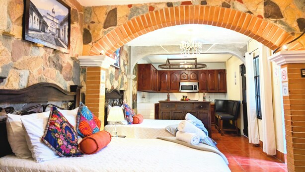 Ein Hotelzimmer mit Steinwand, einem Bett mit bunten Kissen, einer Chandelüster und einem durch einen Bogen sichtbaren Küchenbereich.