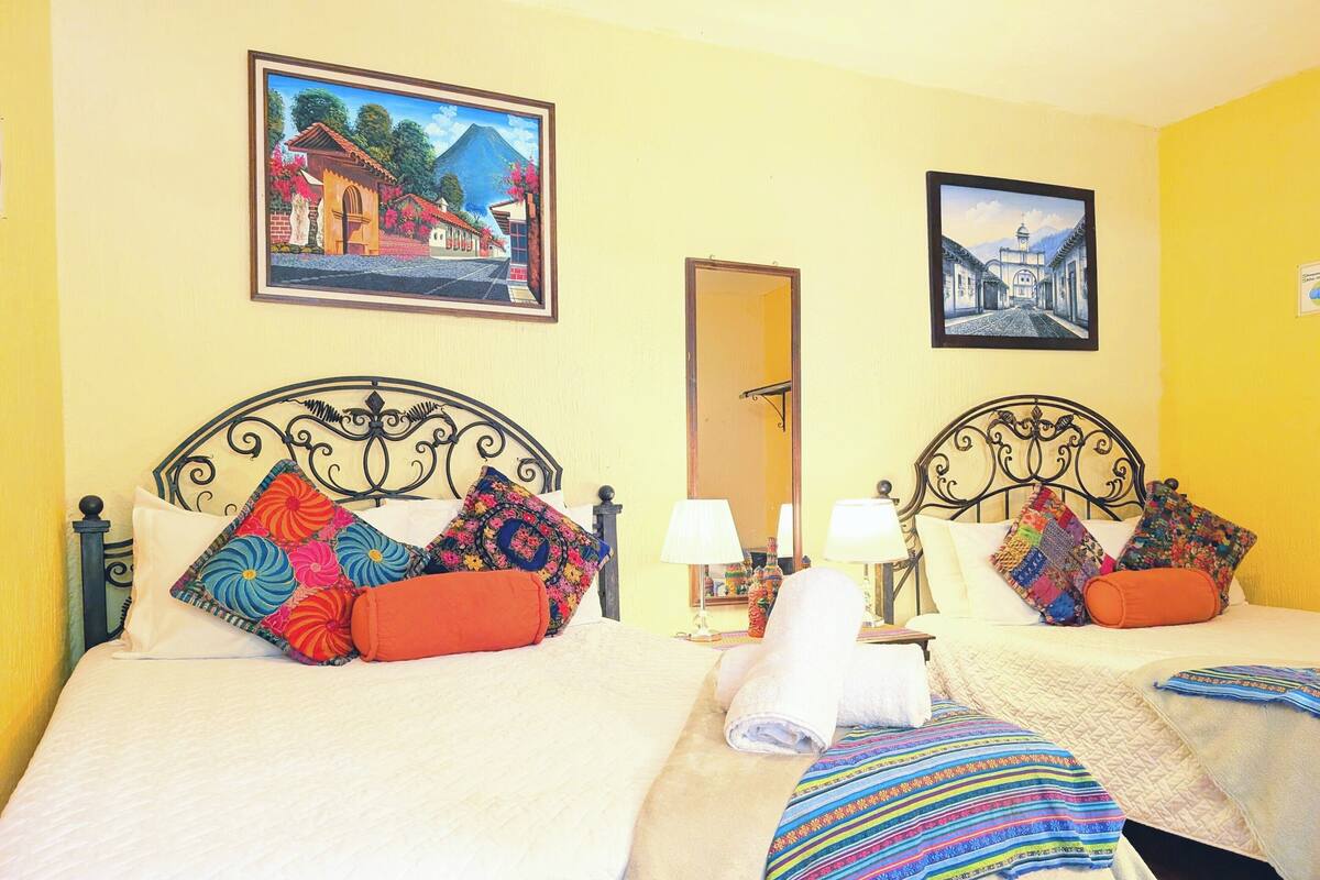 Une chambre d’hôtel avec deux lits, chacun doté d’une tête de lit, d’oreillers et d’une couverture. Il y a deux tableaux accrochés aux murs et un miroir sur la porte.