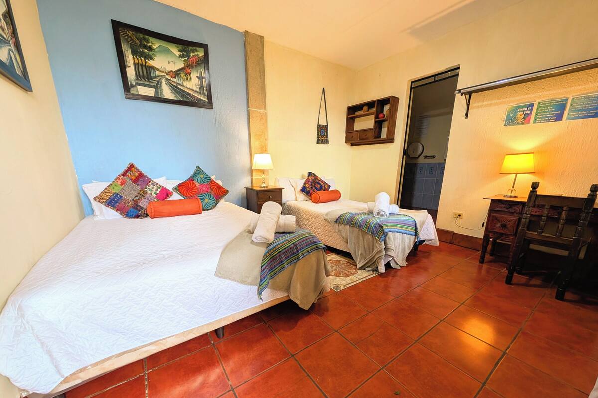 Une chambre d’hôtel avec deux lits, un petit bureau, une chaise, une étagère fixée au mur et une porte donnant sur une salle de bain.