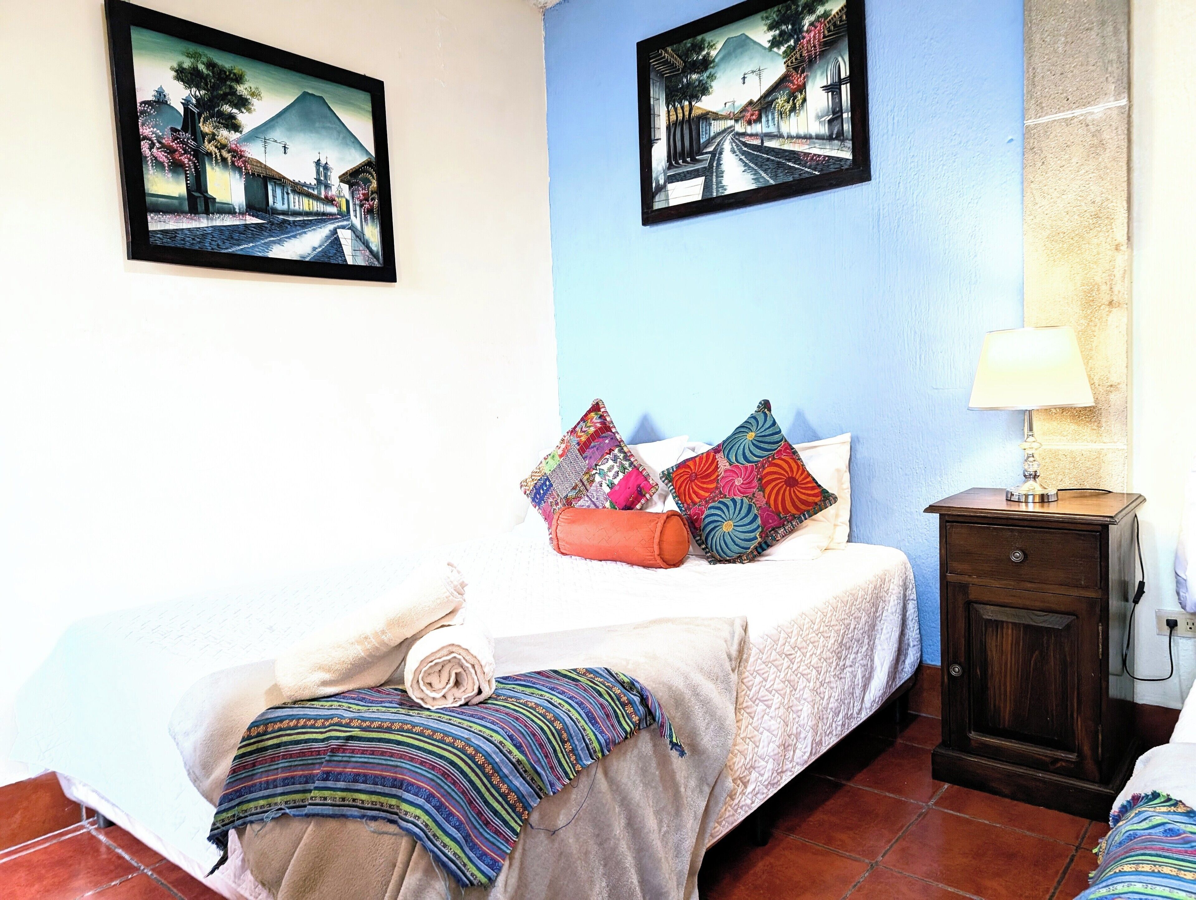 Een netjes opgemaakt bed met kleurrijke kussens, een klein nachtkastje met een lamp, en twee ingelijste kunstwerken aan de muur.