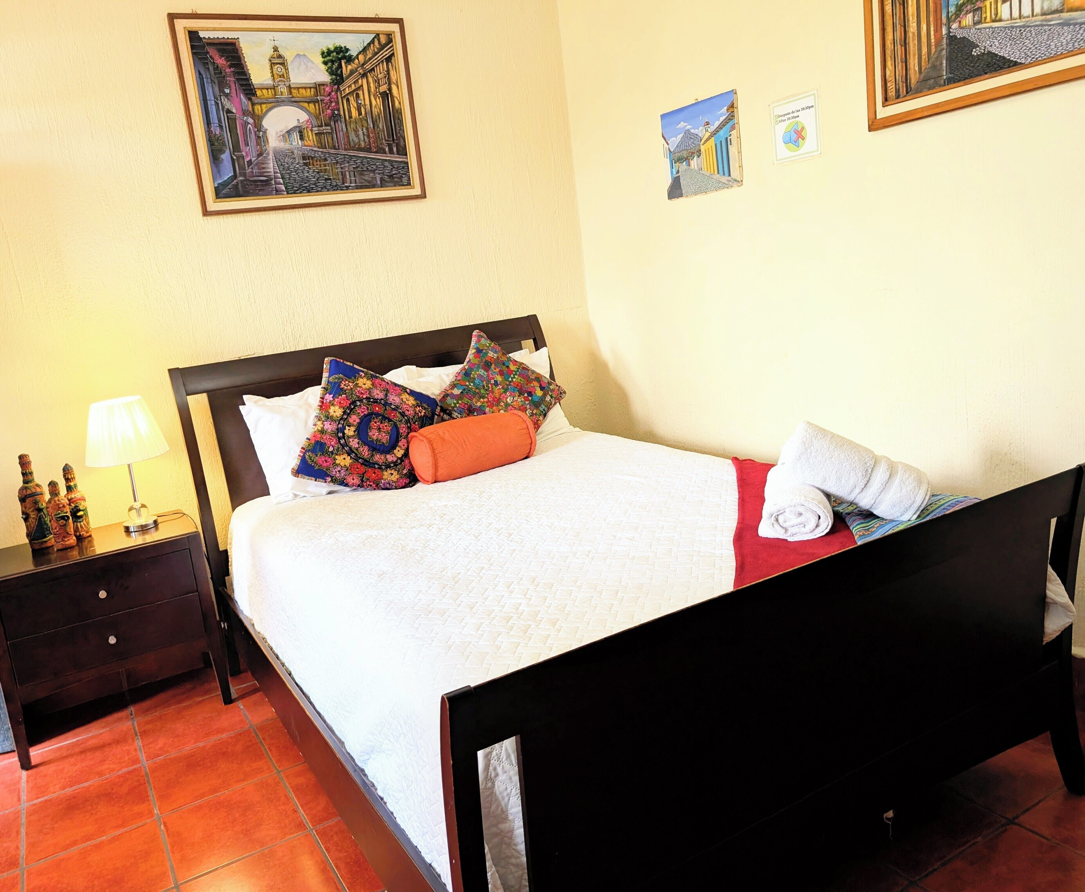 Una cama bien hecha con almohadas coloridas, una toalla blanca y una manta roja. Hay una mesita de noche con una lámpara y botellas decorativas. La habitación tiene suelo de baldosas y un cuadro enmarcado en la pared.