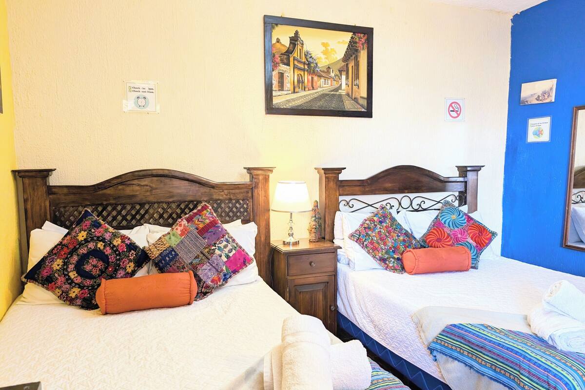 Deux lits avec des couvertures et des oreillers aux couleurs vives, un tableau accroché au mur, une lampe sur une table de chevet et un mur bleu.