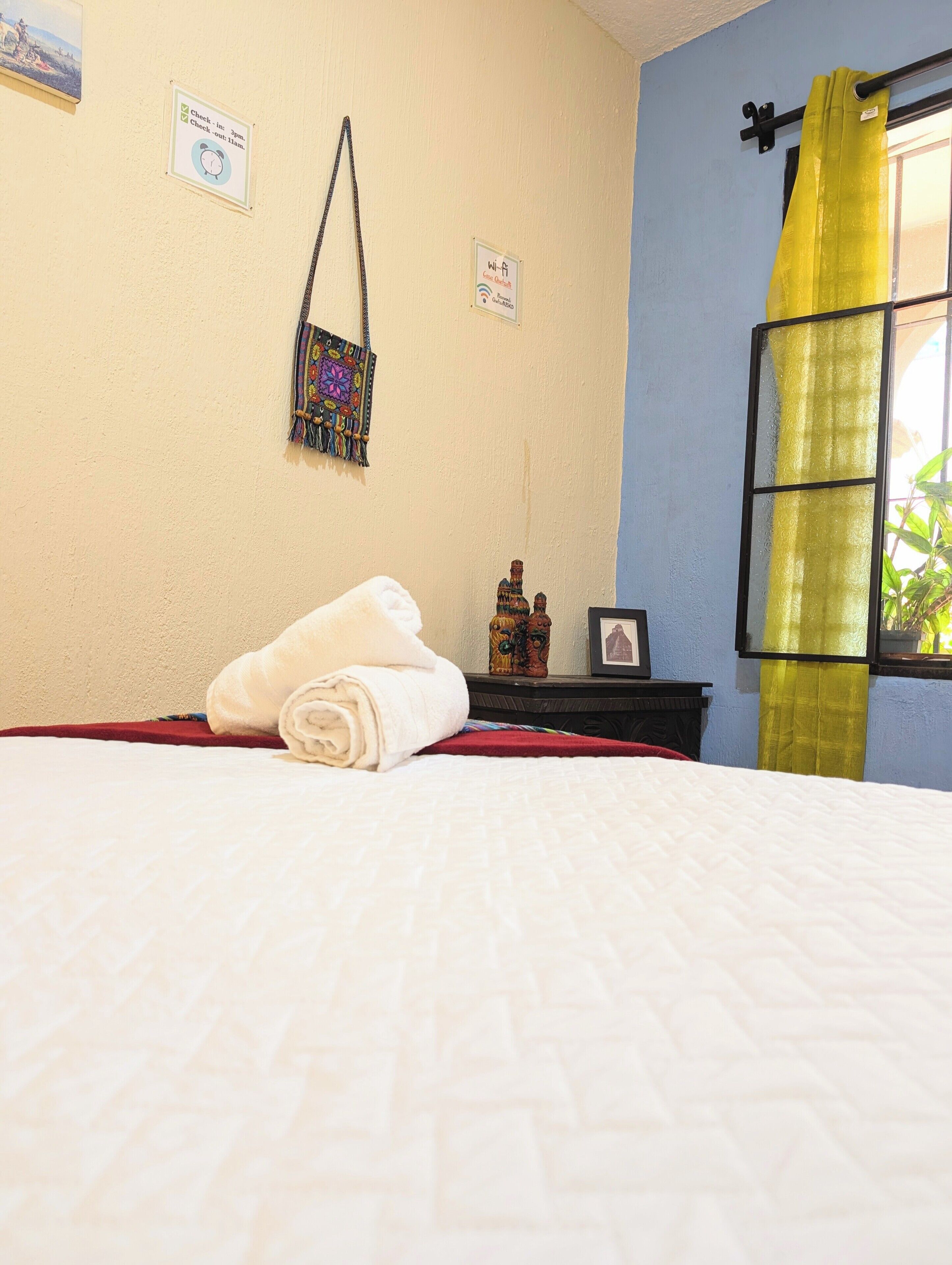 A neatly made bed with a rolled white towel, a small decorative item on a nightstand, and a window with yellow curtains.