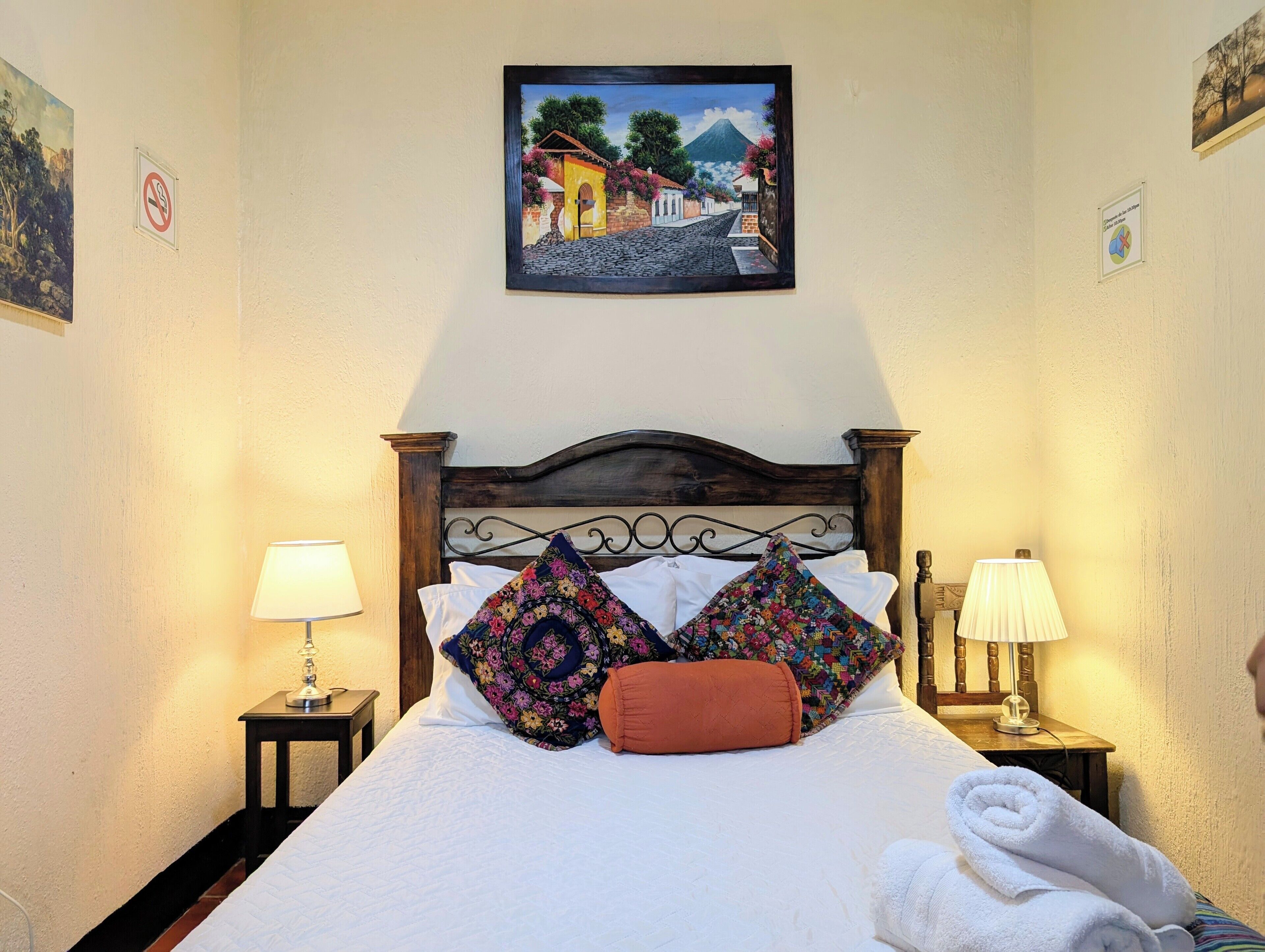 A neatly made bed with floral pillows, a wooden headboard, and a painting of a street scene above it.