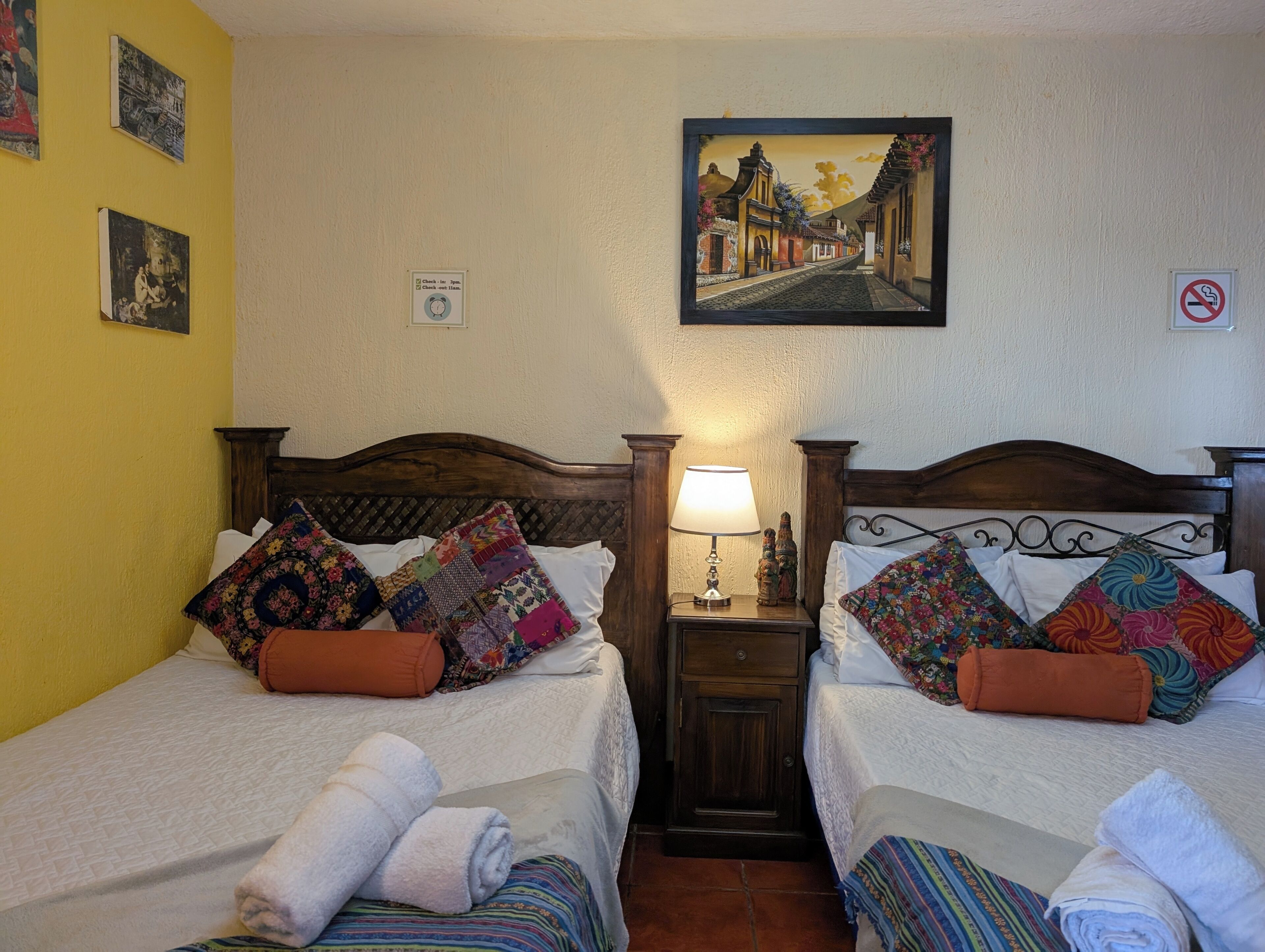 A hotel room with two beds, each with colorful quilts and pillows, a nightstand with a lamp, and a painting on the wall.