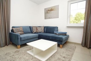 Ein Wohnzimmer mit einem Ecksofa, einem weißen Couchtisch, einem Gemälde an der Wand und einem Fenster mit Vorhängen.