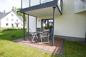 Eine überdachte Terrasse mit Tisch und Stühlen, eine Rasenfläche und ein Gebäude im Hintergrund.