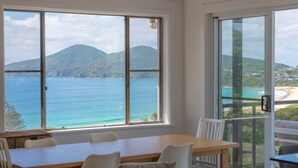 Een kamer met een groot raam dat uitzicht biedt op een strand en bergen. Er staat een eettafel met stoelen in de kamer.