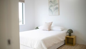 Een minimalistische slaapkamer met een bed, een nachtkastje, een wandlamp en een raam.