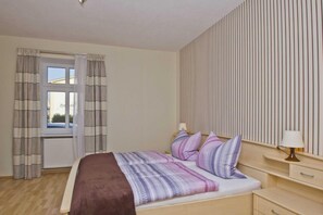 A bedroom with a bed, curtains, a window, a radiator, a bedside table with a lamp, and a wall with vertical lines. - F-1009 Sonnenhut - Ferienwohnung Sonnenhut F-1009 (Baabe)