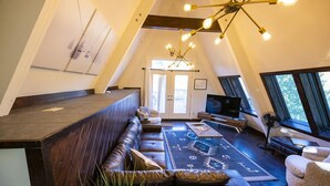 Een ruime woonruimte in loft-stijl met een donker houten bank, een sofa en een bijzettafeltje. Achterin staat een televisie en een ventilator, en een groot raam biedt uitzicht op de omgeving.