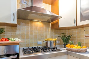 A kitchen with a gas stove, a pot of food, a cutting board with vegetables, and a basket of fruit. - Domos Pintadas Cottage Rosa (Olbia)