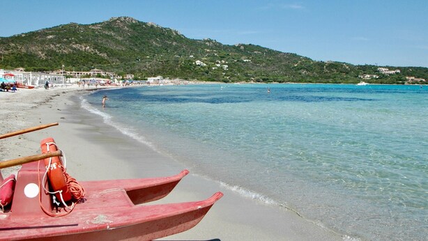 A beach with clear turquoise water, a sandy shore, and a row of red boats. - Domos Pintadas Cottage Rosa (Olbia)