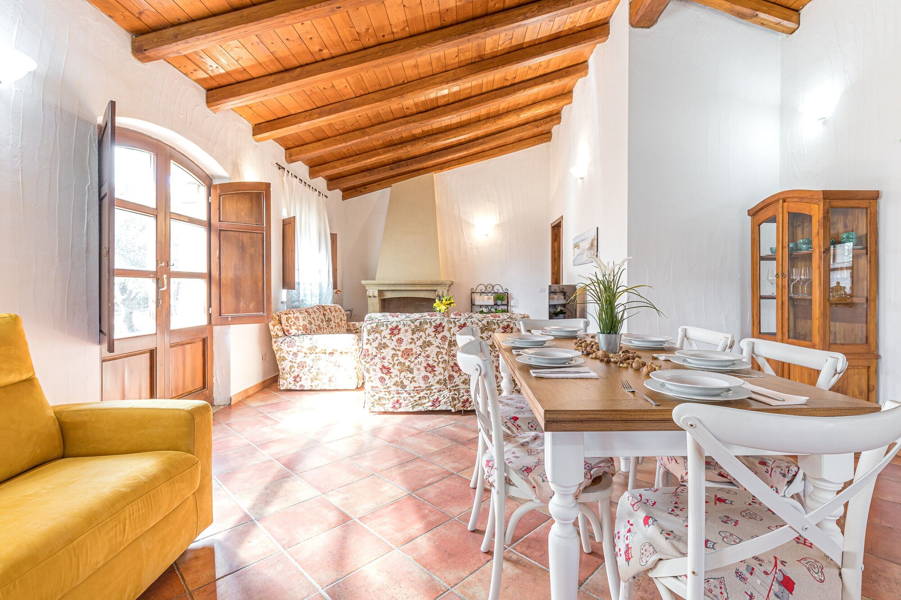 Un comedor con una mesa de madera preparada para cuatro comensales, sillas blancas, un sillón amarillo y un sofá floral.