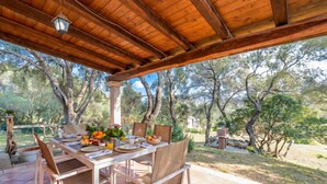 An outdoor dining area with a wooden pergola, a table set with plates, bowls, and a fruit basket. - Domos Pintadas Cottage Rosa (Olbia)