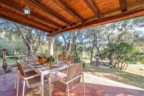 An outdoor dining area with a wooden pergola, a table set with plates, bowls, and a fruit basket. - Domos Pintadas Cottage Rosa (Olbia)