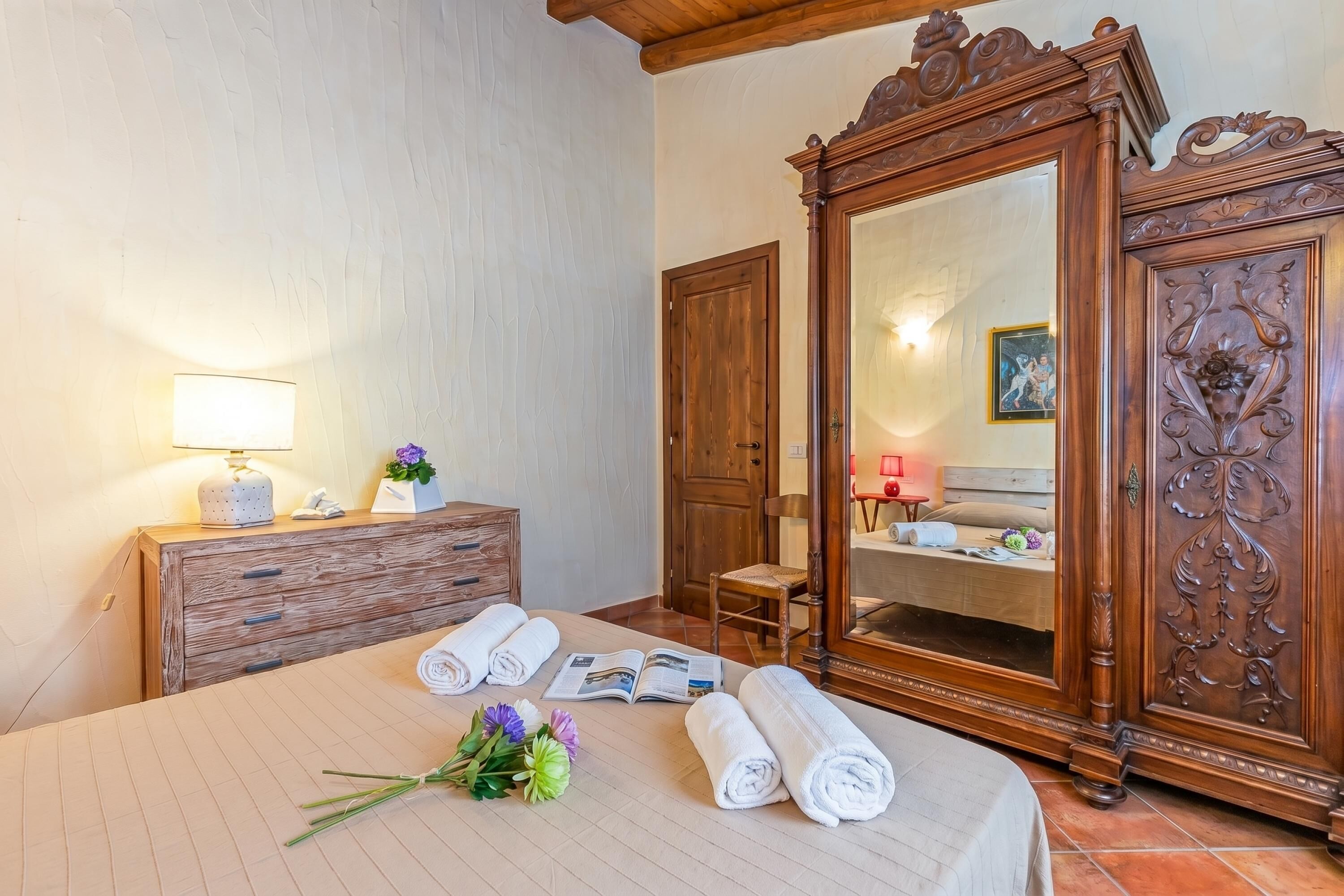 Une chambre d’hôtel avec un lit, une commode en bois, un grand miroir et une table de chevet avec une lampe et des fleurs.