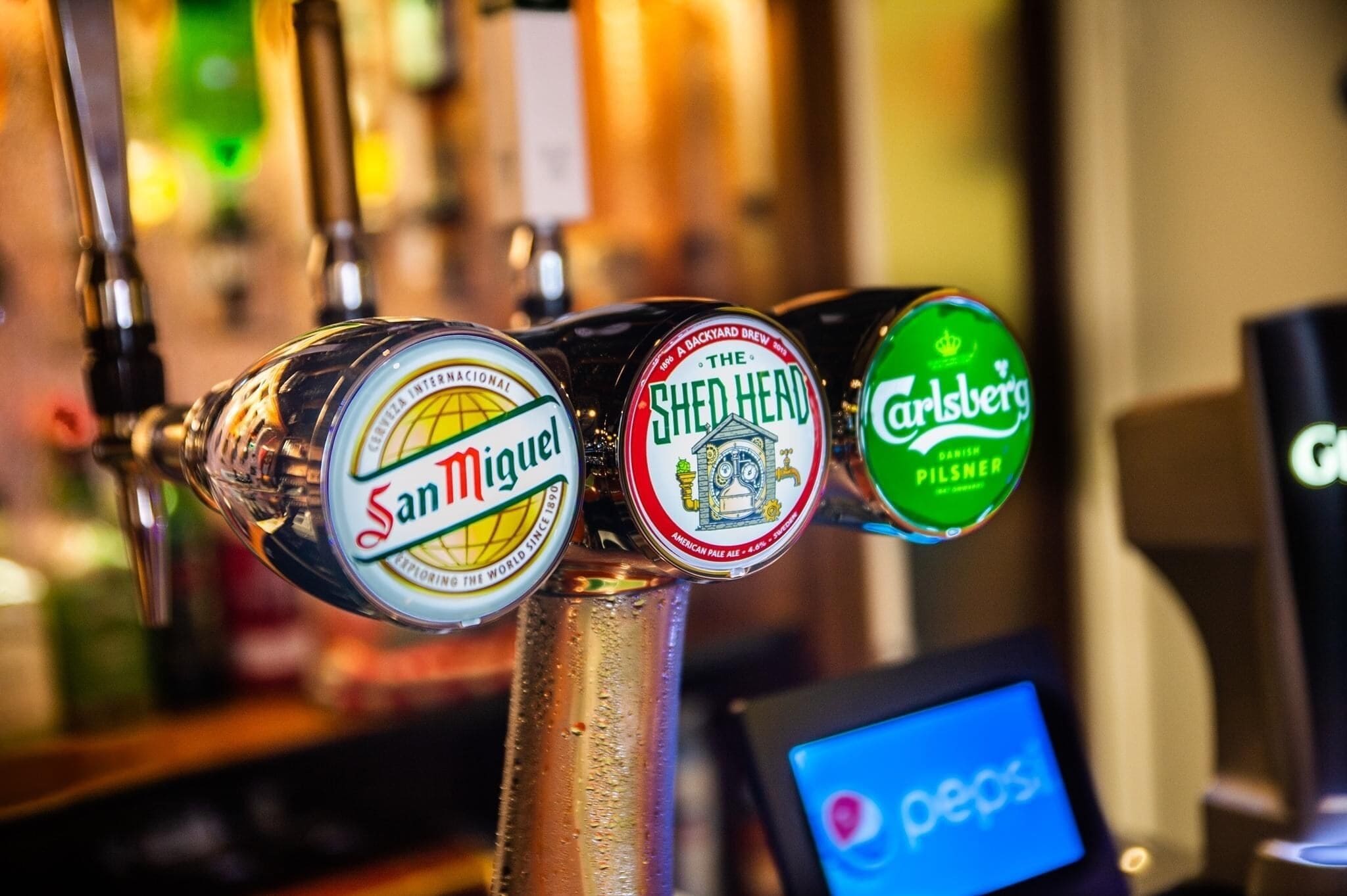 Ein Wasserhahn mit drei Bieretiketten: San Miguel, The Shed Head und Carlsberg.