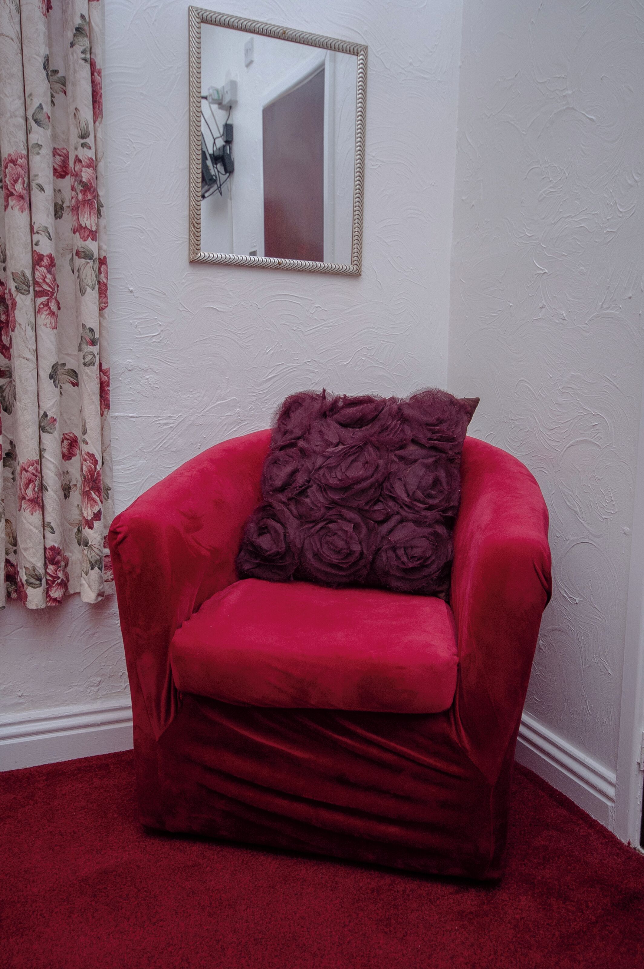 A red velvet armchair with a floral throw pillow, positioned in front of a white wall with a mirror and floral curtains.