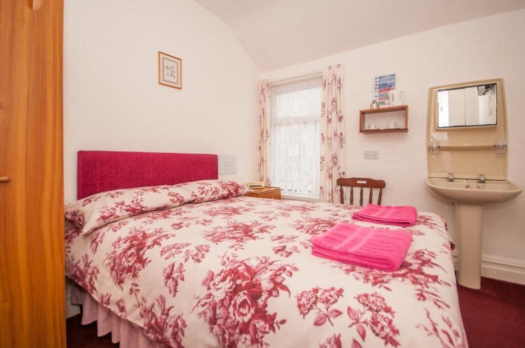 Une chambre à coucher avec un lit, une table de chevet, une fenêtre avec des rideaux, un miroir et un lavabo.