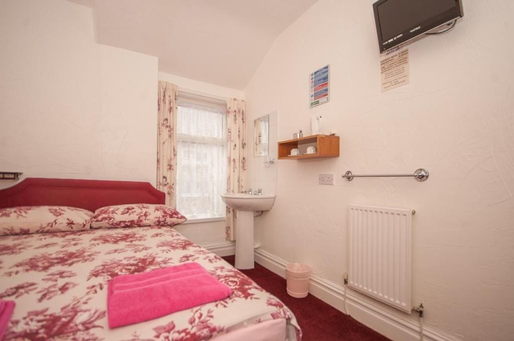 A bedroom with a bed, a window with curtains, a bedside table, a radiator, and a mounted television.