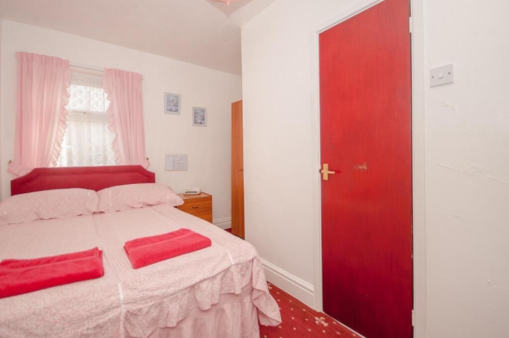 Un dormitorio con cama, mesita de noche, puerta roja y ventana con cortinas.