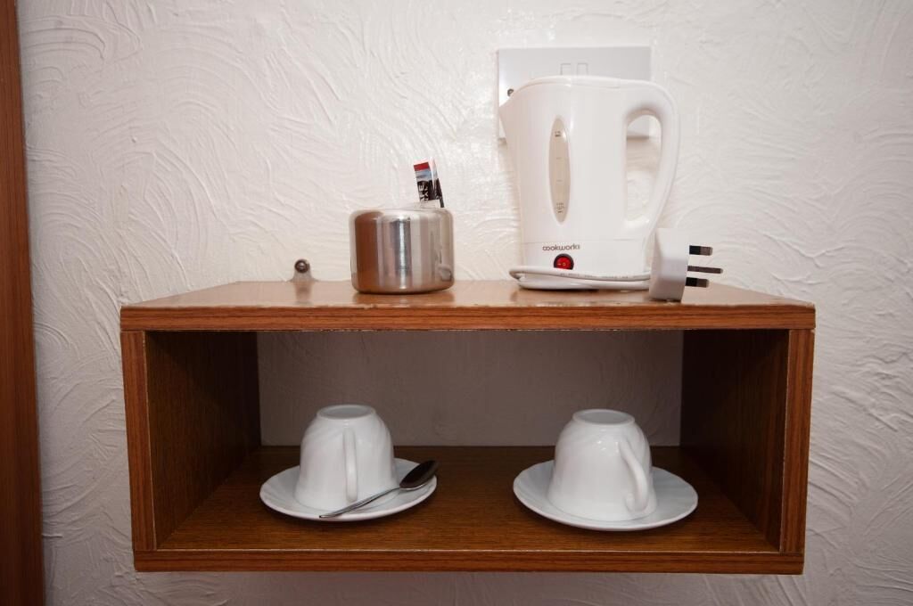 A wooden shelf with a white electric kettle and a container, and two white cups with saucers below.