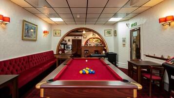 A billiards room with a pool table, red seating, and a bar area in the background.