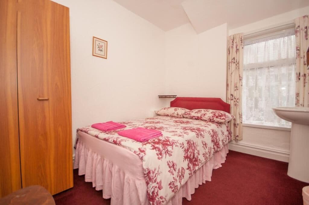 A bedroom with a bed, a wooden wardrobe, a window with curtains, and a wall-mounted shelf.