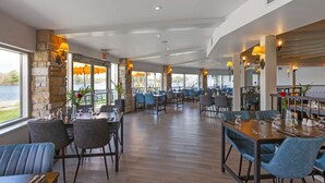 A spacious restaurant with high ceilings, stone walls, and large windows overlooking a body of water. Tables are set with blue chairs and centerpieces. - Christie's Mill Inn & Spa, BW Premier Collection (Georgian Bay)